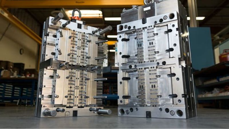 Two open metal injection molds stand amid machinery and tools in an industrial setting.