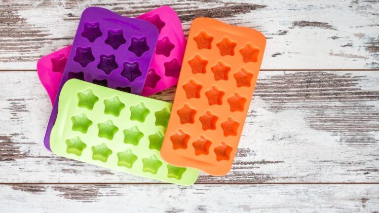 Quatre bacs à glaçons en silicone en forme d'étoile en violet, orange, vert et rose sur une surface en bois blanc.